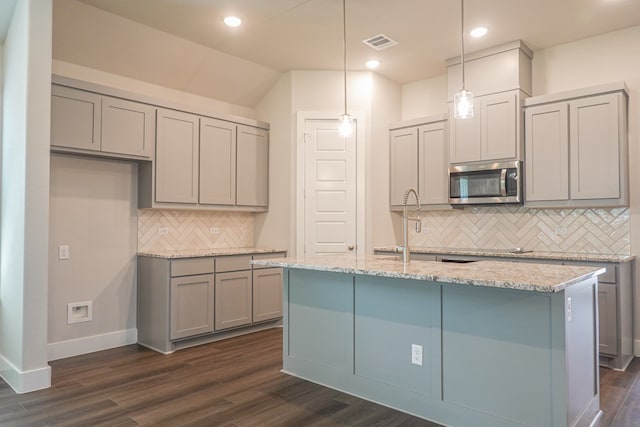 kitchen featuring dark hardwood / wood-style flooring, decorative backsplash, hanging light fixtures, a kitchen island with sink, and light stone counters