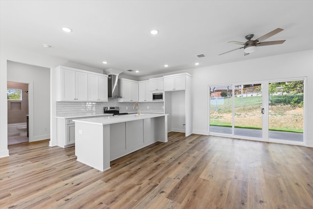 kitchen with light hardwood / wood-style floors, white cabinetry, wall chimney exhaust hood, stainless steel appliances, and a center island with sink