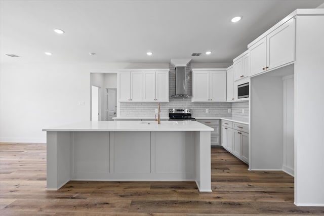 kitchen featuring dark wood-type flooring, wall chimney range hood, an island with sink, and stainless steel appliances