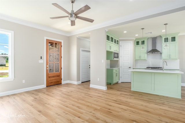 kitchen with light hardwood / wood-style flooring, ceiling fan, hanging light fixtures, a center island with sink, and wall chimney range hood