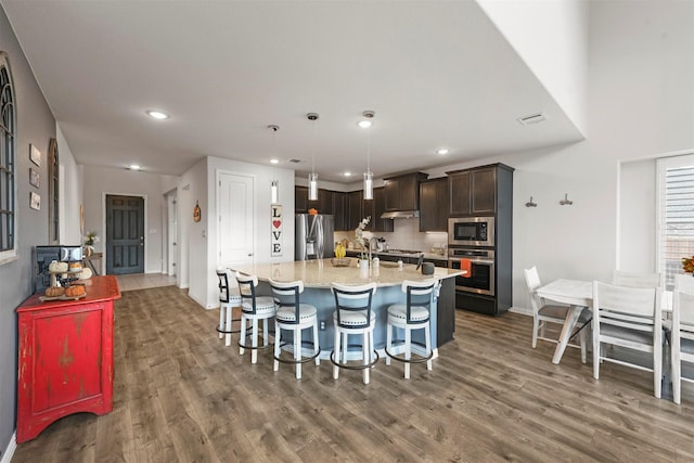 kitchen featuring pendant lighting, stainless steel appliances, dark brown cabinetry, an island with sink, and dark hardwood / wood-style flooring