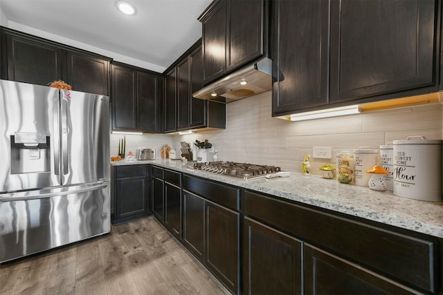 kitchen with appliances with stainless steel finishes, tasteful backsplash, dark brown cabinetry, light stone countertops, and light wood-type flooring