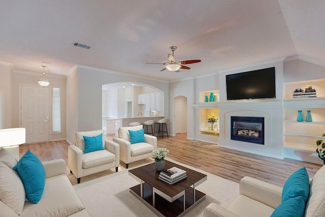 living room featuring ceiling fan, built in shelves, light hardwood / wood-style floors, crown molding, and vaulted ceiling