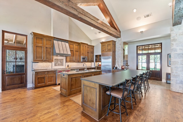 kitchen with custom range hood, light hardwood / wood-style floors, an island with sink, a breakfast bar area, and built in appliances