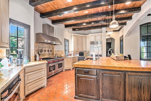 kitchen with wall chimney range hood, an island with sink, decorative backsplash, butcher block counters, and double oven range