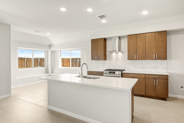 kitchen with stainless steel range, sink, wall chimney range hood, backsplash, and an island with sink