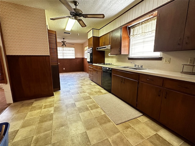 kitchen with wall oven, ceiling fan, sink, black dishwasher, and wood walls