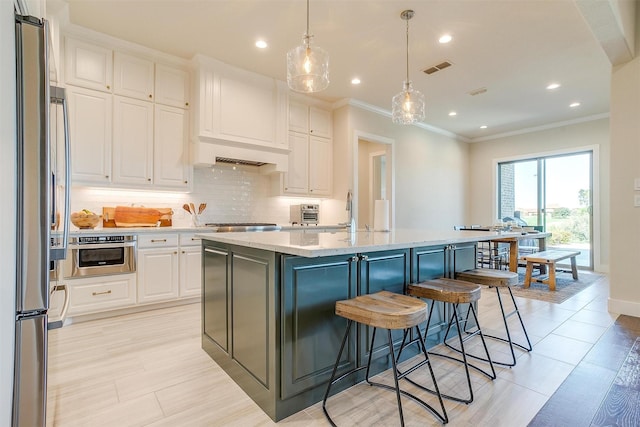 kitchen featuring appliances with stainless steel finishes, an island with sink, light stone counters, white cabinetry, and decorative light fixtures