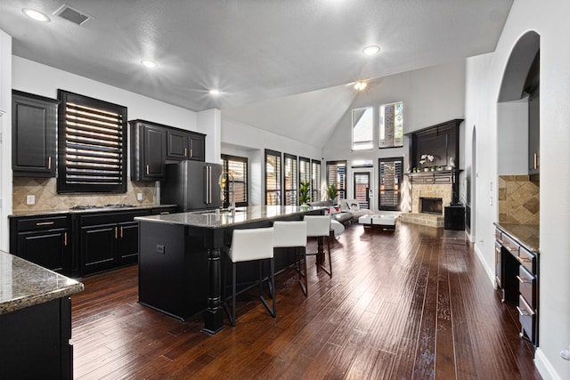 kitchen with high end refrigerator, stone counters, a fireplace, a kitchen island with sink, and dark hardwood / wood-style flooring