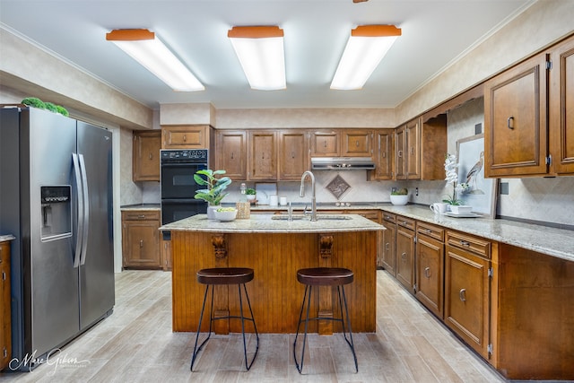 kitchen featuring a breakfast bar area, black double oven, an island with sink, light stone countertops, and stainless steel refrigerator with ice dispenser