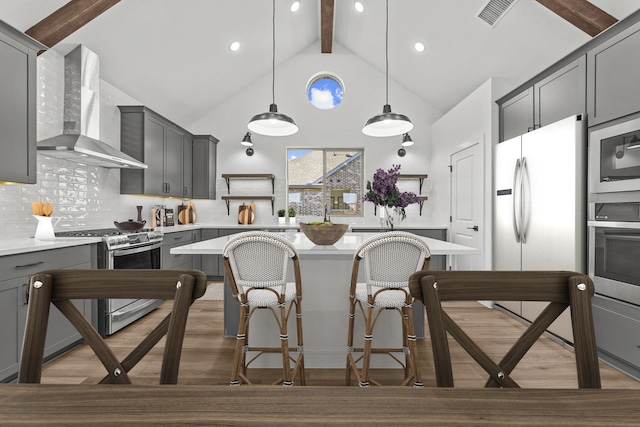 kitchen with wall chimney exhaust hood, gray cabinetry, stainless steel appliances, beamed ceiling, and hanging light fixtures