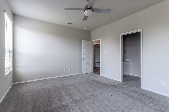 unfurnished bedroom featuring dark colored carpet, a spacious closet, ceiling fan, and ensuite bath