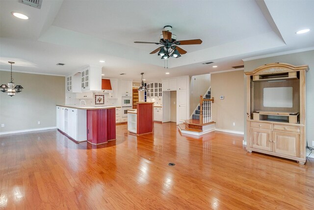 unfurnished living room with ceiling fan with notable chandelier, a raised ceiling, ornamental molding, and light hardwood / wood-style flooring