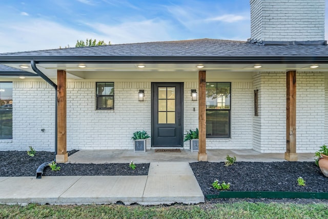 view of exterior entry featuring a porch, brick siding, and a shingled roof