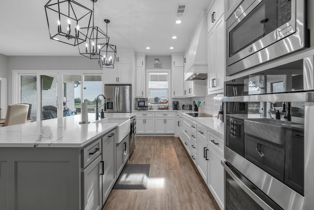 kitchen featuring stainless steel appliances, custom range hood, white cabinetry, a sink, and an island with sink