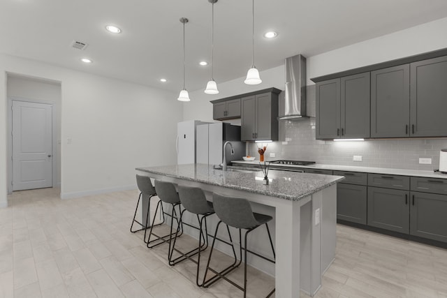 kitchen featuring decorative backsplash, light stone countertops, an island with sink, hanging light fixtures, and wall chimney range hood