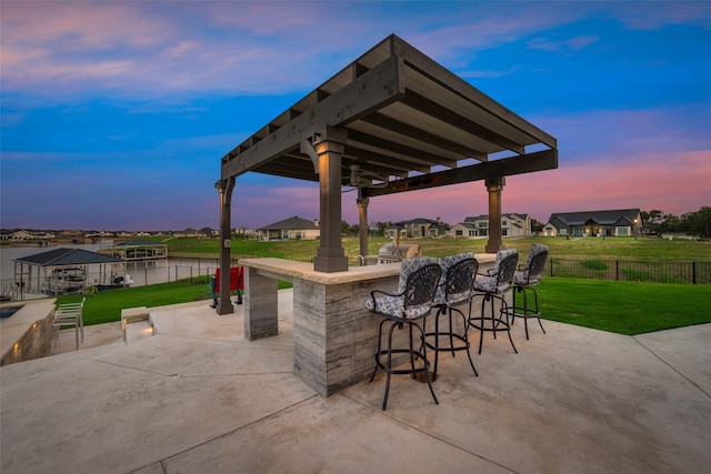 patio terrace at dusk with a water view, a lawn, and a bar