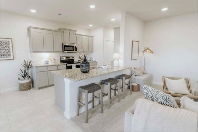 kitchen featuring appliances with stainless steel finishes, gray cabinets, light stone counters, a breakfast bar, and a center island with sink