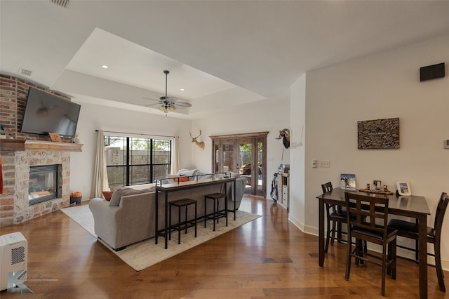 living room with ceiling fan, brick wall, a stone fireplace, a tray ceiling, and hardwood / wood-style floors