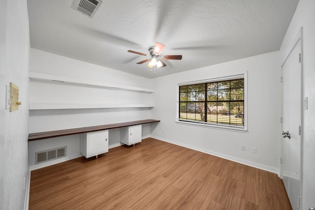 unfurnished office with ceiling fan, built in desk, a textured ceiling, and light hardwood / wood-style flooring