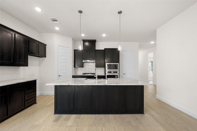 kitchen with hanging light fixtures, a center island with sink, appliances with stainless steel finishes, light stone countertops, and light hardwood / wood-style floors