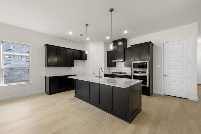 kitchen with tasteful backsplash, light stone counters, a kitchen island with sink, light hardwood / wood-style flooring, and stainless steel appliances