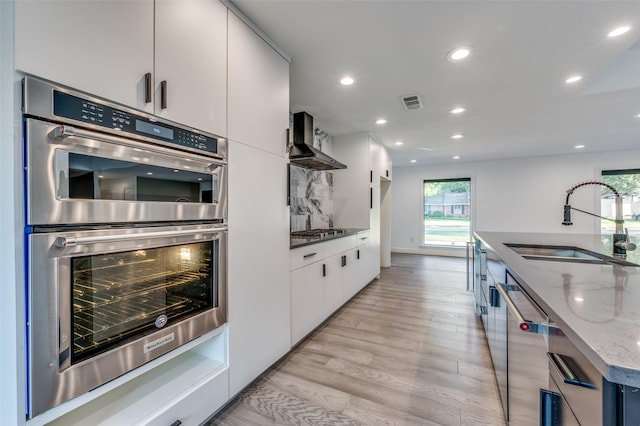 kitchen featuring sink, white cabinetry, appliances with stainless steel finishes, light stone countertops, and wall chimney range hood