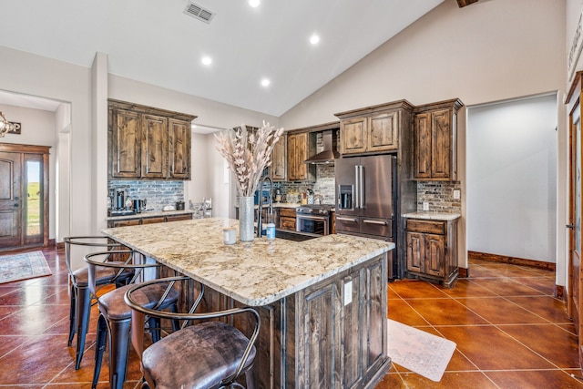 kitchen with appliances with stainless steel finishes, light stone counters, backsplash, a center island with sink, and wall chimney range hood