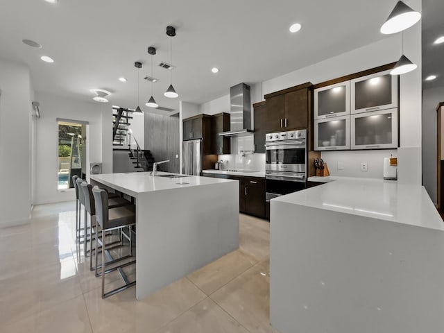 kitchen with stainless steel appliances, wall chimney exhaust hood, dark brown cabinets, sink, and light tile patterned floors