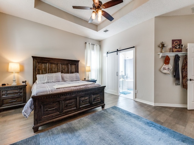 bedroom with a tray ceiling, a barn door, dark hardwood / wood-style flooring, and ceiling fan