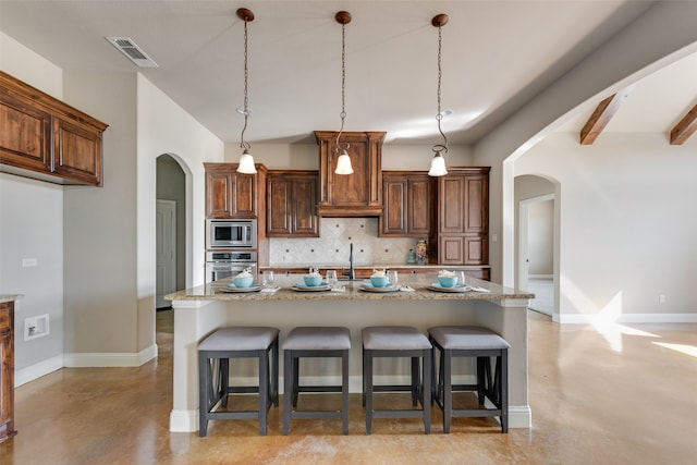 kitchen featuring decorative backsplash, a kitchen island with sink, light stone countertops, pendant lighting, and stainless steel appliances