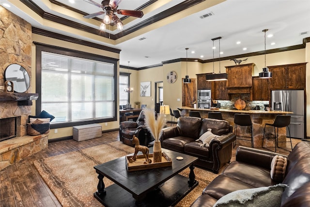 living room with ceiling fan, a raised ceiling, a stone fireplace, hardwood / wood-style floors, and crown molding