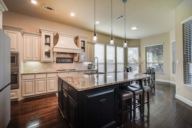 kitchen with light stone countertops, sink, premium range hood, pendant lighting, and a kitchen island with sink