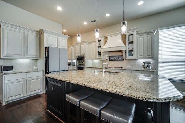 kitchen with tasteful backsplash, custom exhaust hood, a large island with sink, decorative light fixtures, and fridge