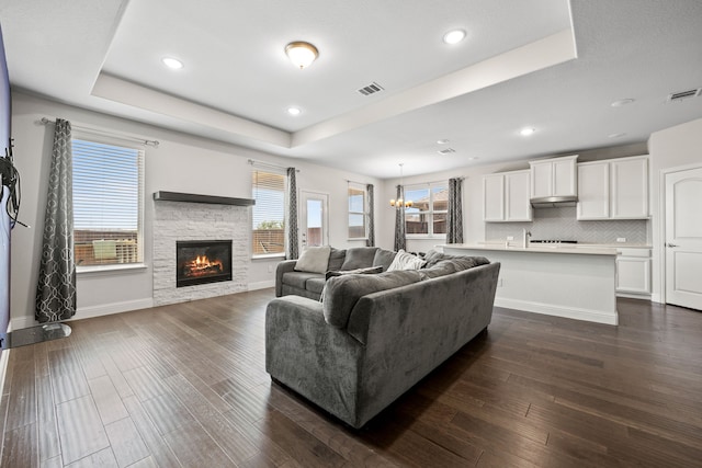 living room with a tray ceiling, dark hardwood / wood-style floors, an inviting chandelier, and a stone fireplace