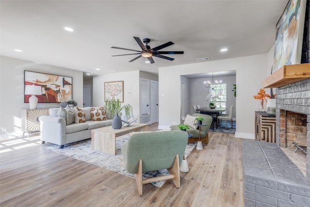 living room with ceiling fan with notable chandelier, a brick fireplace, and light hardwood / wood-style floors