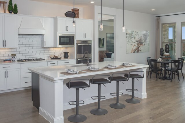 kitchen with appliances with stainless steel finishes, decorative light fixtures, white cabinets, dark wood-type flooring, and a center island with sink