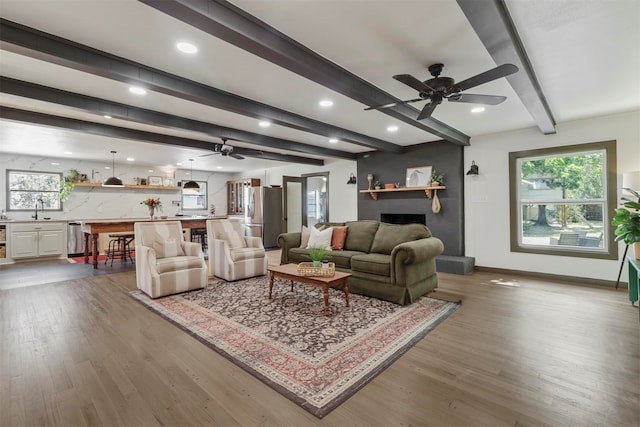 living room featuring beam ceiling, a large fireplace, sink, wood-type flooring, and ceiling fan