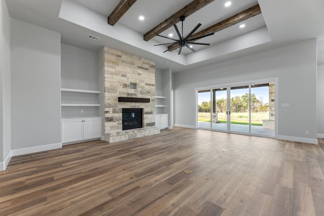 unfurnished living room featuring built in shelves, a stone fireplace, wood-type flooring, beamed ceiling, and ceiling fan