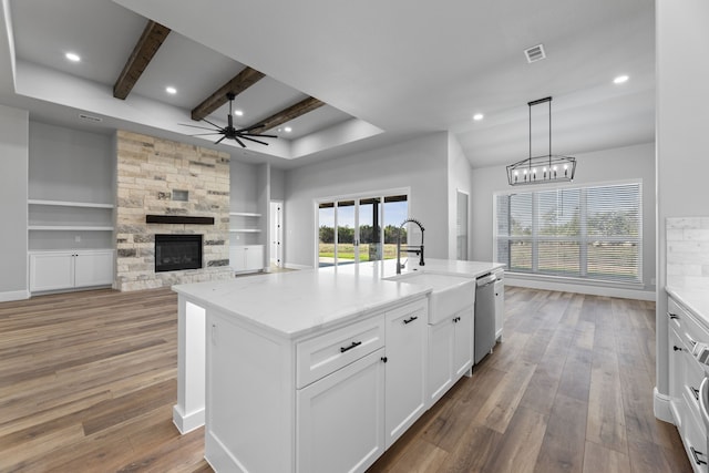 kitchen featuring a stone fireplace, decorative light fixtures, white cabinets, hardwood / wood-style flooring, and a center island with sink