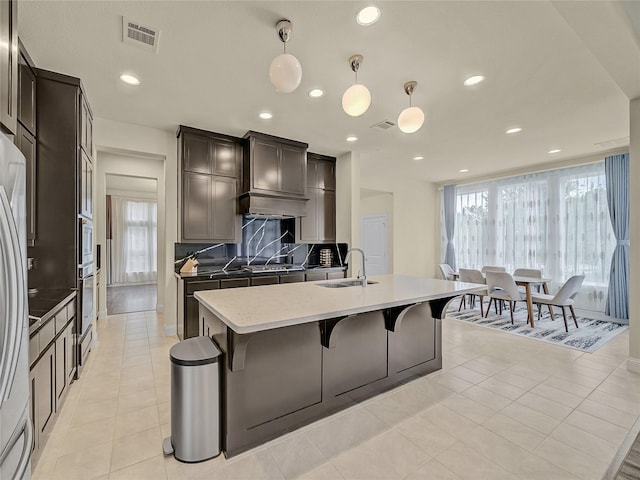 kitchen featuring visible vents, hanging light fixtures, decorative backsplash, a sink, and an island with sink