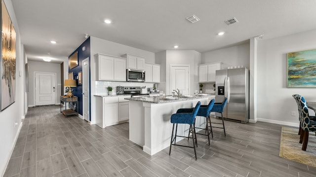 kitchen with appliances with stainless steel finishes, backsplash, light stone counters, a center island with sink, and white cabinets