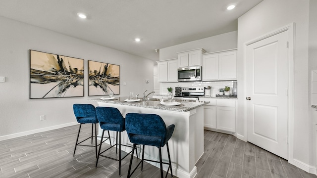 kitchen with backsplash, appliances with stainless steel finishes, light stone counters, an island with sink, and white cabinetry