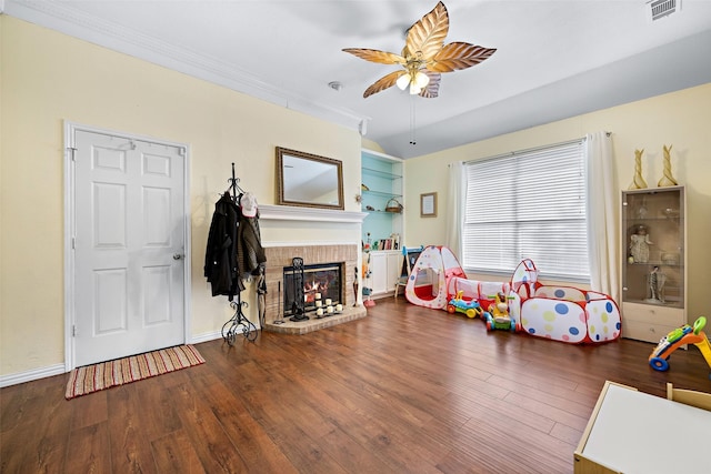 game room featuring visible vents, ceiling fan, dark wood-style flooring, crown molding, and a brick fireplace