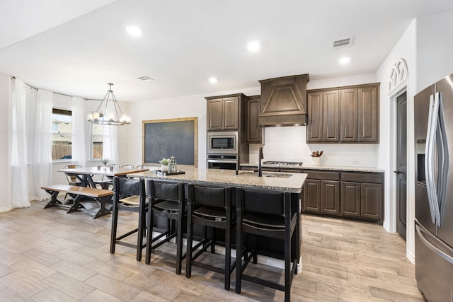 kitchen with a center island with sink, sink, light stone countertops, appliances with stainless steel finishes, and custom range hood