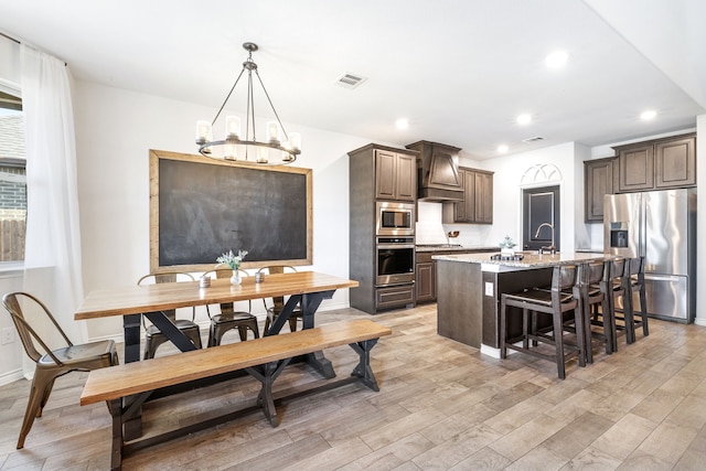 kitchen featuring appliances with stainless steel finishes, custom exhaust hood, dark brown cabinetry, a center island with sink, and light hardwood / wood-style floors