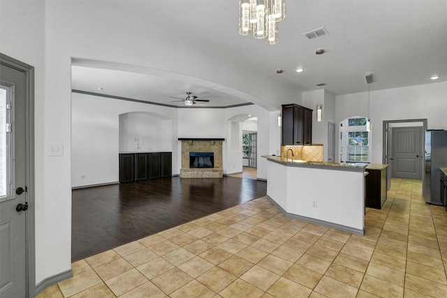 kitchen with a fireplace, light hardwood / wood-style floors, ceiling fan with notable chandelier, tasteful backsplash, and stainless steel refrigerator