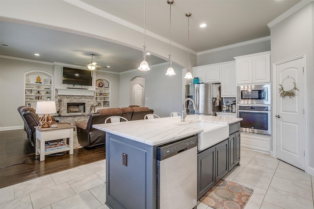 kitchen featuring a stone fireplace, appliances with stainless steel finishes, a kitchen island with sink, ceiling fan, and white cabinets