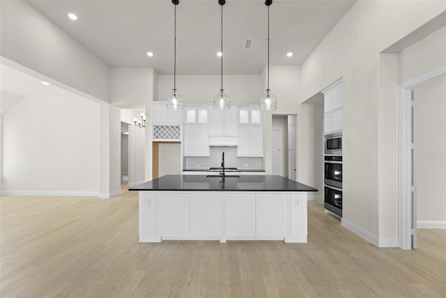 kitchen with decorative backsplash, a kitchen island with sink, decorative light fixtures, white cabinets, and stainless steel microwave