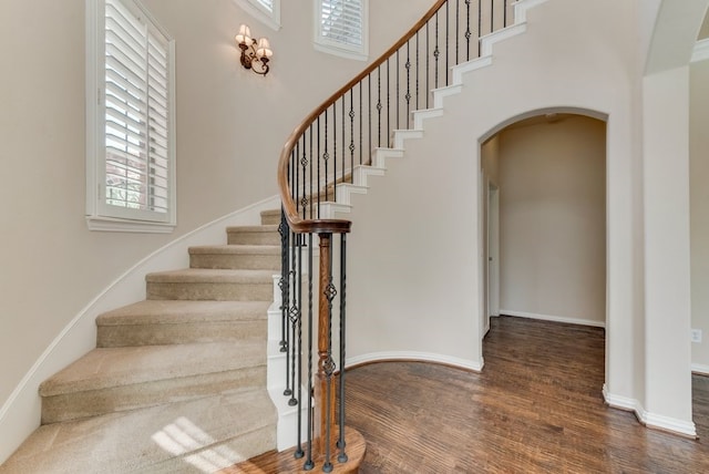 staircase featuring arched walkways, wood finished floors, a towering ceiling, and baseboards